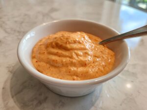 A close-up of thick, creamy orange burger sauce speckled with red spice, served in a small white bowl with a spoon.