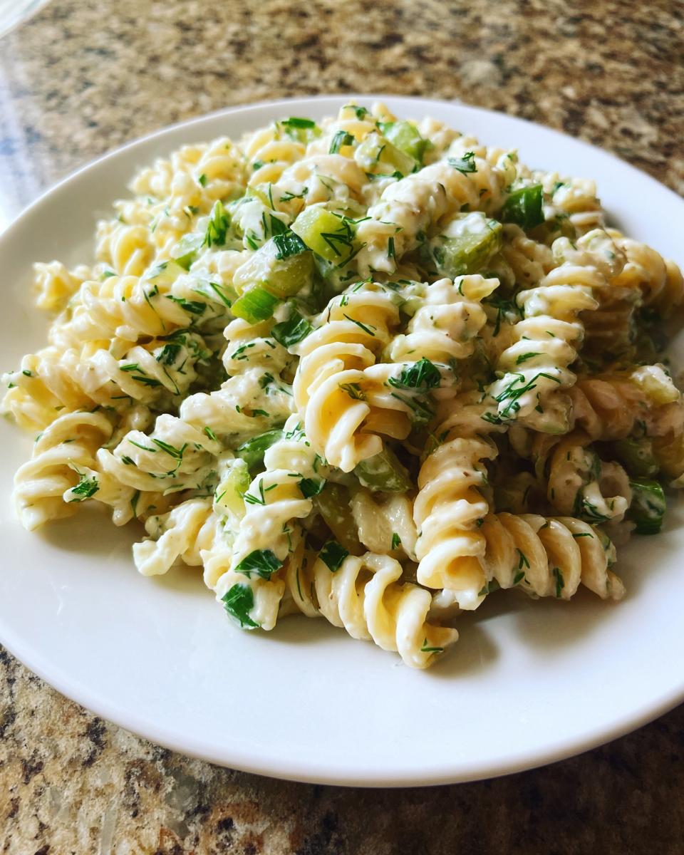 A close-up of creamy pickle pasta salad made with rotini pasta, chopped pickles, and fresh dill on a white plate.