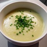 A close-up of creamy potato leek soup served in a white bowl and garnished generously with chopped fresh chives.