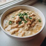 A close-up of a white bowl filled with rich, creamy white chicken chili, topped with shredded chicken, white beans, and fresh cilantro.