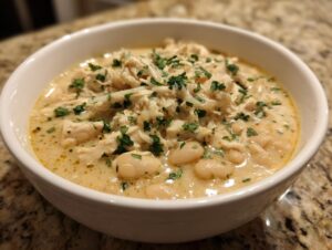 A close-up of a white bowl filled with creamy white chicken chili, featuring shredded chicken, white beans, and a parsley garnish.