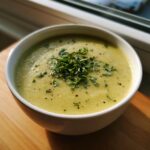 A close-up of a bowl of creamy, light green zucchini soup topped generously with fresh chopped herbs and black pepper.
