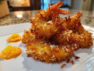 Close-up of perfectly fried, crispy coconut shrimp served on a white plate with orange dipping sauce.