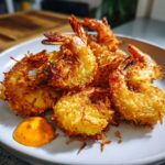 A close-up of perfectly fried, golden brown coconut shrimp piled on a white plate next to a dollop of orange dipping sauce.