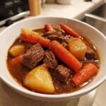 A close-up of a hearty bowl of crockpot beef stew featuring tender beef chunks, bright carrots, and potatoes in a rich brown gravy.