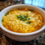 A close-up of a white bowl filled with golden broth and delicate ribbons of egg drop soup, garnished with chopped green onions.