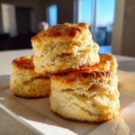 Close-up of several tall, flaky buttermilk biscuits stacked on a white plate, golden brown and perfectly risen.