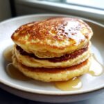 A close-up stack of three fluffy almond flour pancakes drizzled generously with golden syrup.