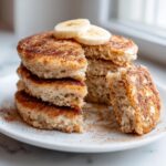 A stack of fluffy banana oatmeal pancakes dusted with cinnamon, topped with banana slices, with one piece cut out.