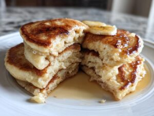 A fluffy stack of banana pancakes cut in half, drizzled with syrup and topped with banana slices.