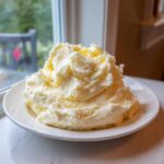 A large, fluffy mound of rich, creamy mashed potatoes served on a white plate near a window.