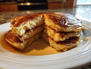 A stack of fluffy sourdough discard pancakes, cut open to show the texture and drizzled with maple syrup.