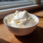 A white bowl filled with freshly whipped, fluffy vanilla buttercream frosting, sitting on a wooden surface near a window.