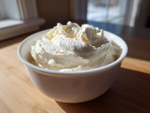 A white bowl filled with freshly whipped, fluffy vanilla buttercream frosting sitting on a wooden surface.