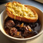 Close-up of a bowl of dark, rich french onion beef stew topped with a slice of toasted bread covered in melted, bubbly cheese.