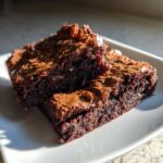 Two rich, fudgy brownies with shiny, crackly tops stacked slightly on a white plate.