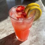 A tall glass filled with bright pink lemonade, ice, sliced strawberries, and lemon garnishes.