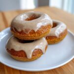 Three golden baked pumpkin donuts stacked on a white plate, topped with a thick, speckled brown sugar glaze.