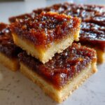 Close-up of two stacked, gooey pecan pie bars showing a thick, dark caramel topping over a shortbread crust.