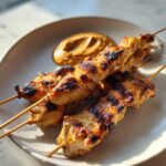 Four grilled chicken satay skewers served on a white plate next to a dollop of creamy peanut sauce.