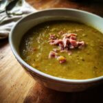 Close-up of a thick, green pea soup in a white bowl, garnished with shredded ham.