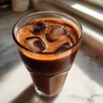 Close-up of a tall glass filled with iced chocolate milk, featuring a rich, frothy top and several ice cubes.