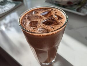 Close-up of a tall glass filled with iced chocolate milk, topped with foam and ice cubes, glistening in the sunlight.
