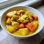 Close-up of Jamaican curry chicken served in a white bowl with chunks of potato and bright orange carrots in a rich yellow sauce.