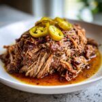 A close-up of shredded Mississippi pot roast served in a white bowl with rich juices and topped with sliced pickled jalapeños.