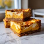 Three stacked squares of keto pumpkin cheesecake bars showing a creamy orange swirl filling and dark crust.