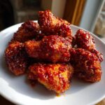 Close-up of crispy Korean fried chicken recipe wings coated in a thick, glossy, deep red spicy sauce with sesame seeds.