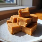 A stack of creamy, light brown maple fudge squares resting on a speckled white plate near a window.