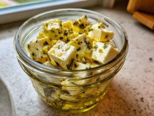 Close-up of cubed marinated cheese soaking in olive oil with herbs and garlic in a small glass jar.