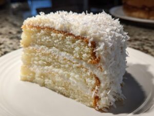 A moist slice of layered coconut cake, frosted and heavily topped with shredded coconut flakes.