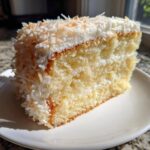 Close-up of a moist slice of coconut cake with white frosting and toasted coconut flakes on top.