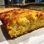A close-up of a golden brown slice of moist cream cheese corn casserole studded with whole corn kernels.