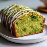 A close-up of a slice of moist pistachio bread loaf topped with white icing drizzle and chopped pistachios.