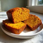 Three thick slices of vibrant orange, moist pumpkin bread stacked on a white plate.