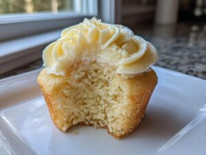 Close-up of a moist vanilla cupcake with a bite taken out, showing the fluffy interior and swirl of vanilla frosting.