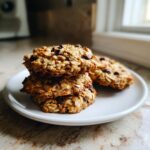 A stack of three oatmeal chocolate chip breakfast cookie on a white plate near a window.