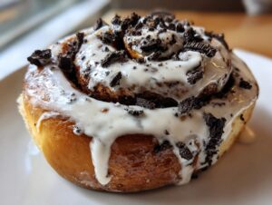 A close-up shot of a single, freshly baked Oreo cinnamon rolls covered in thick white icing and crushed Oreo pieces.