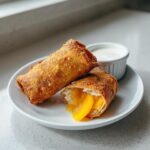 Two crispy, golden brown peach cobbler egg rolls on a white plate, one cut open showing warm peach filling.