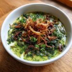 A bowl of vibrant green Persian noodle soup topped generously with crispy fried onions and fresh parsley.