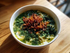 Close-up of a white bowl filled with vibrant green Persian noodle soup, topped with fried onions.