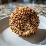 A close-up of a finished pineapple cheese ball completely coated in chopped toasted nuts on a white plate.
