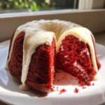A slice cut from a vibrant red velvet bundt cake topped with thick white cream cheese glaze.