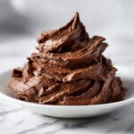 A tall swirl of rich, dark chocolate frosting mounded in a small white bowl on a marble surface.