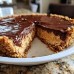 A close-up of a decadent peanut butter pie with a thick peanut butter filling, graham cracker crust, and glossy chocolate ganache topping, one slice removed.
