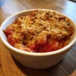 Close-up of a single serving of warm rhubarb crisp with a golden, crumbly topping in a white ramekin.