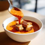 A spoonful of thick, vibrant roasted tomato soup being poured over croutons in a white bowl.